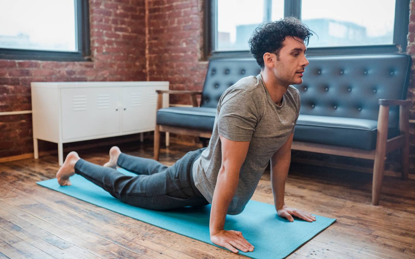 Un joven descalzo hace la posición de la cobra en el living de un departamento, sobre un mat de yoga.