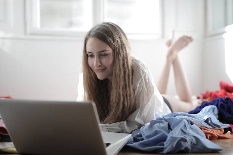 Una mujer joven está acostada en el suelo  mirando una laptop, en medio de ropa tirada.