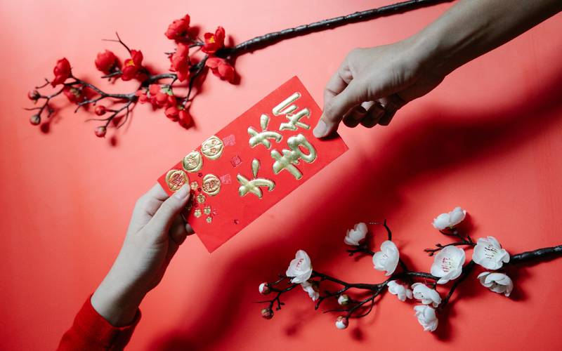 Fondo rojo sobre el cual hay ramas de cerezos en flor y dos manos pasándose una tarjeta con ideogramas chinos.