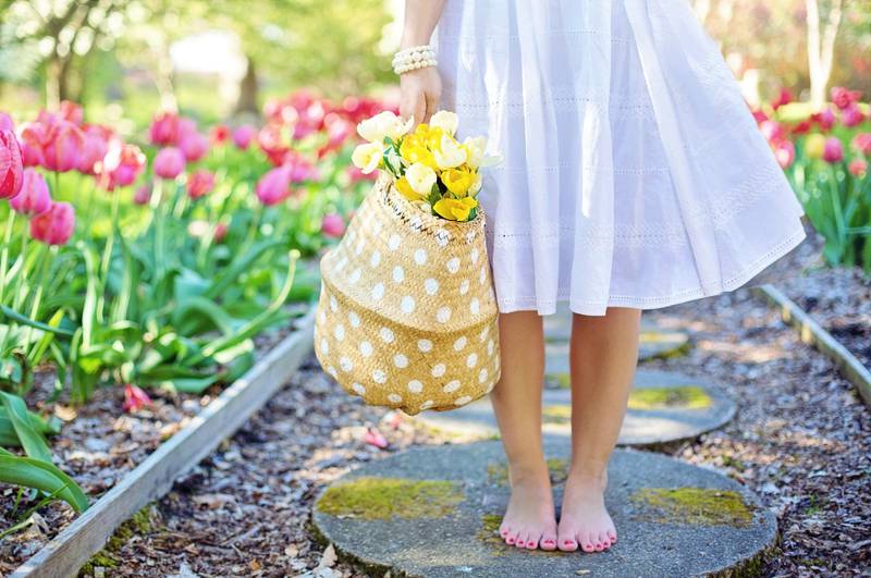 Unas piernas femeninas y juveniles están descalzas en un jardín. La chica lleva una canasta con flores y un vestido blanco.