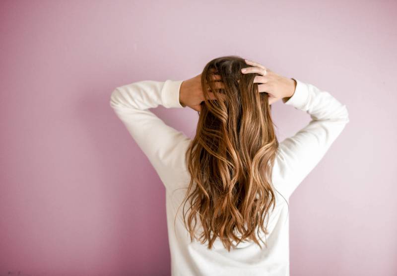 Persona de cabello largo, rizado y rubio oscuro, vista de espalda. Está vestida de blanco y tiene los brazos en la cabeza. El fondo es de color rosa.