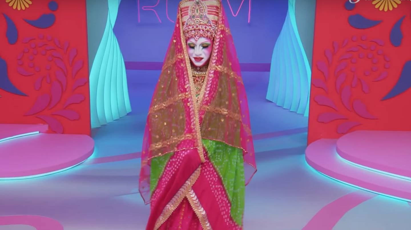 toma de cuerpo entero de drag vestida como personaje de la india, con un tocado alto en tono rojizos y verdosos