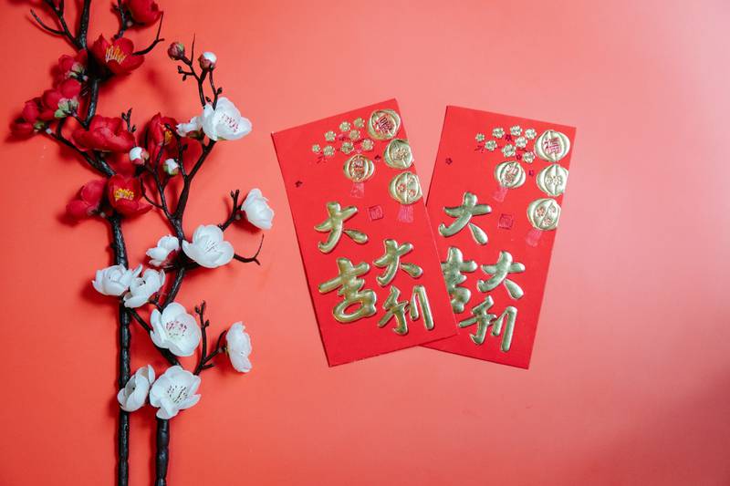 Dos ramas de cerezos en flor acompañan a dos tarjetas con caracteres  y decoraciones chinas. Los cuatro elementos están sobre un fondo rojo pálido.