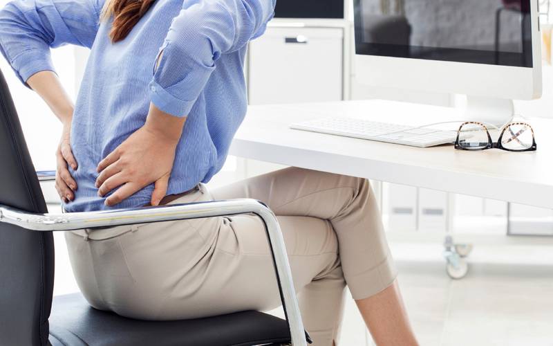 Una mujer sentada frente a un escritorio con las piernas ruzadas, posa sus manos en la espalda en un gesto de dolor