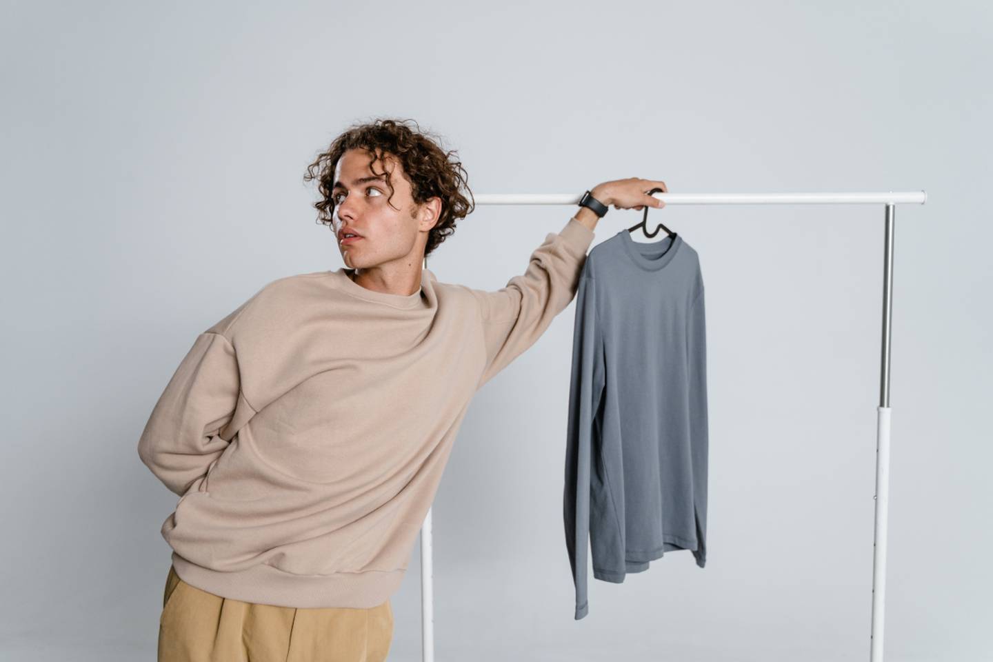 Un hombre joven sostiene un colgador de ropa donde pende sólo una prenda. La otra mano está oculta en su espalda y mira hacia la izquierda.