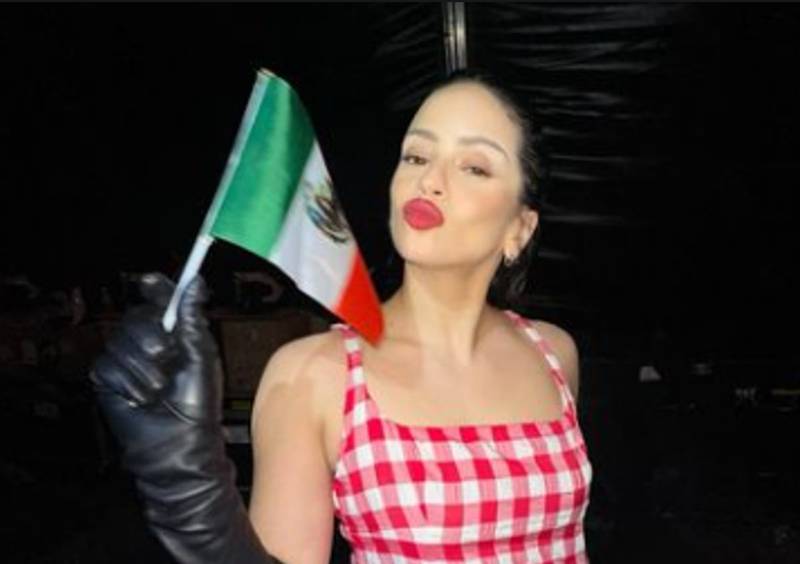 Mujer con vestido a cuadros de tirantes, con tranzas y guantes de cuero negros, con una bandera mexicana en la mano