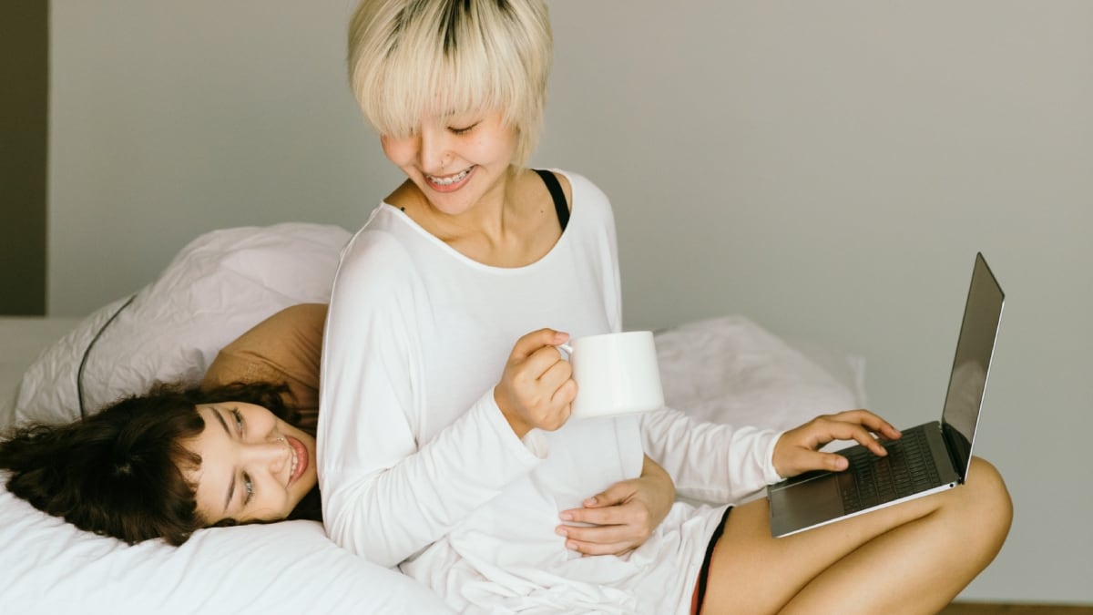 Una pareja descansa sobre una cama. Una de ellas está sentada con una laptop en las piernas y una taza en la mano. Ambas se sonríen cómplices.
