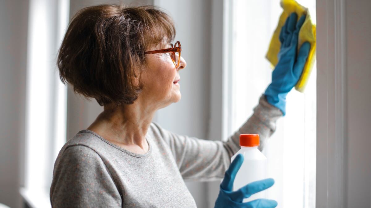 Una mujer de edad avanzada limpia con guantes y un paño amapirro el cristal de una ventala. La mujer lleva lentes.