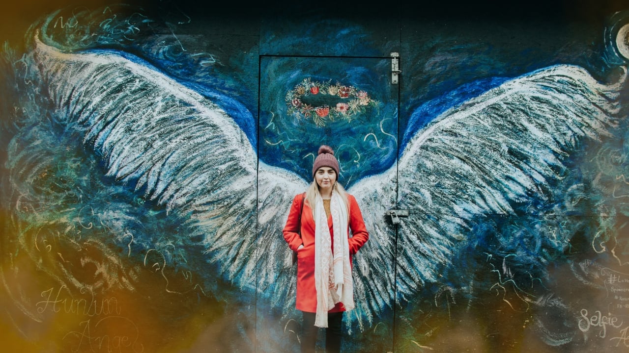Foto de una mujer adelante de un mural de fondo azúl que consiste en un par de alas y una aureola que le dan la apariencia de un ángel. La mujer es rubia y, a juzgar por su atuendo de abrigo, parece una foto exterior en un día frío.
