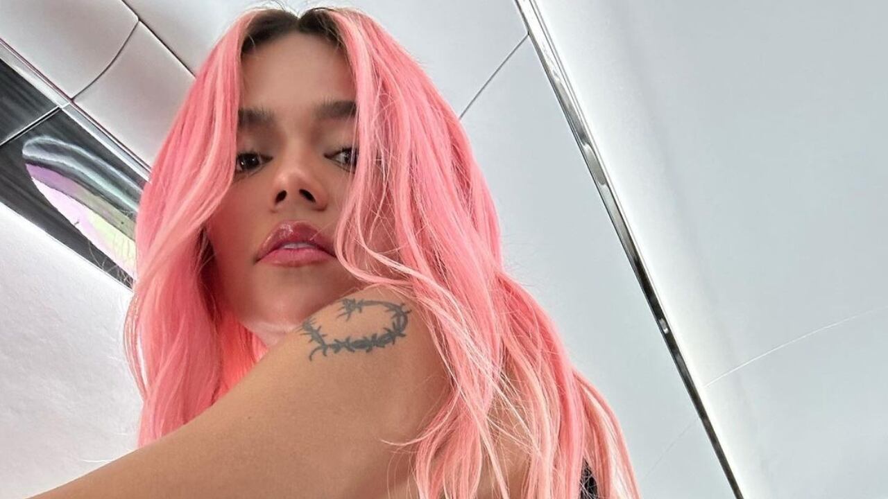 Karol G al interior de un avión, con pelo suelto y rosado en una toma desde abajo. En su hombro izquierdo se puede ver el tatuaje de corazón.