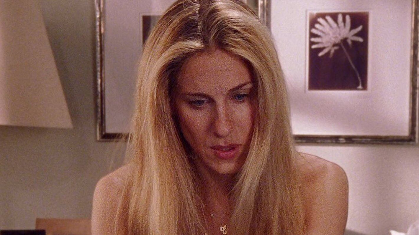 Primero plano a Sarah Jessica Parker, rubia con el pelo peinado al medio y con una cadena de oro que dice "Carrie", en el fondo se ven unos cuadros con una flor dibujada cada uno