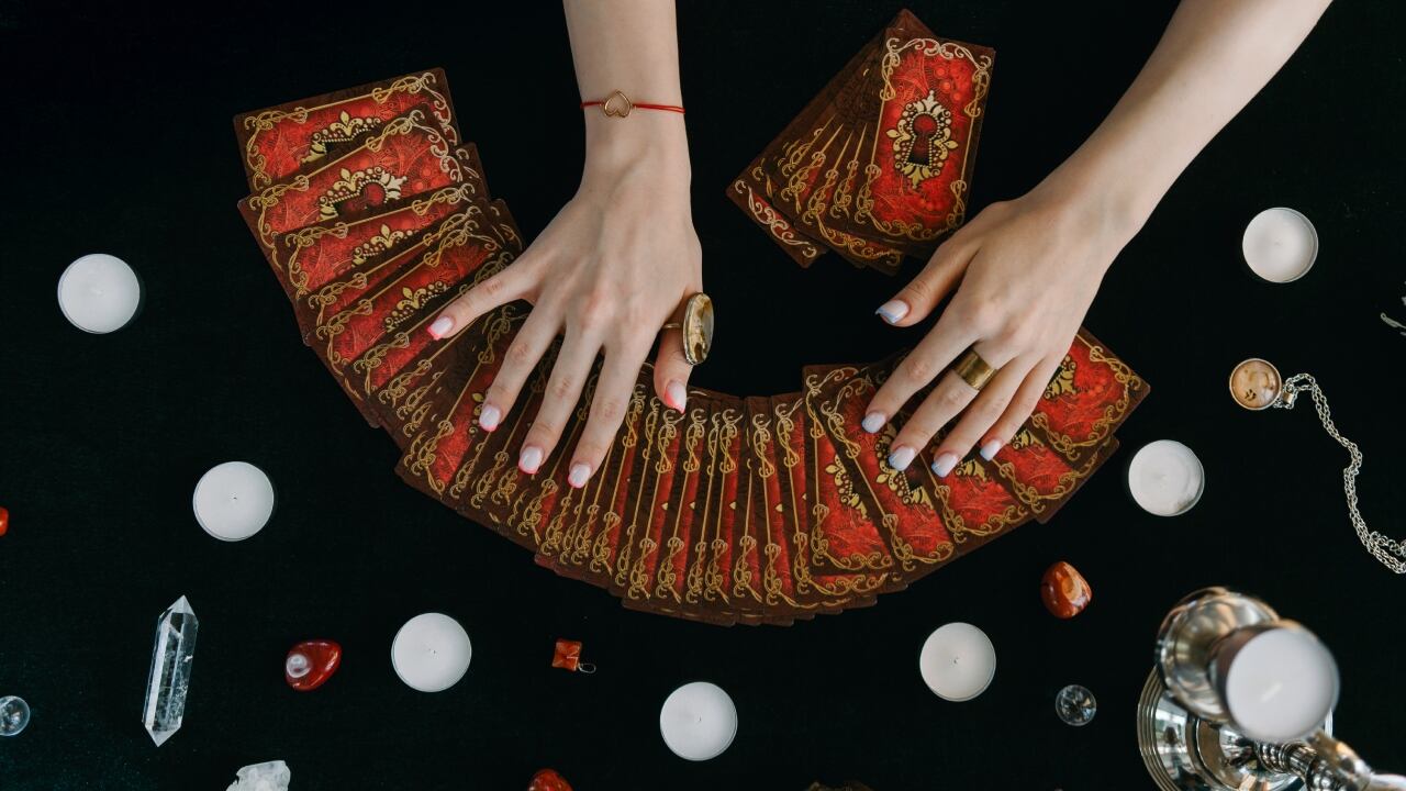 Un par de manos femeninas extendiendo una baraja de tarot con las figuras hacia abajo, sobre un paño negro. Sobre el paño también hay velas y piedras.