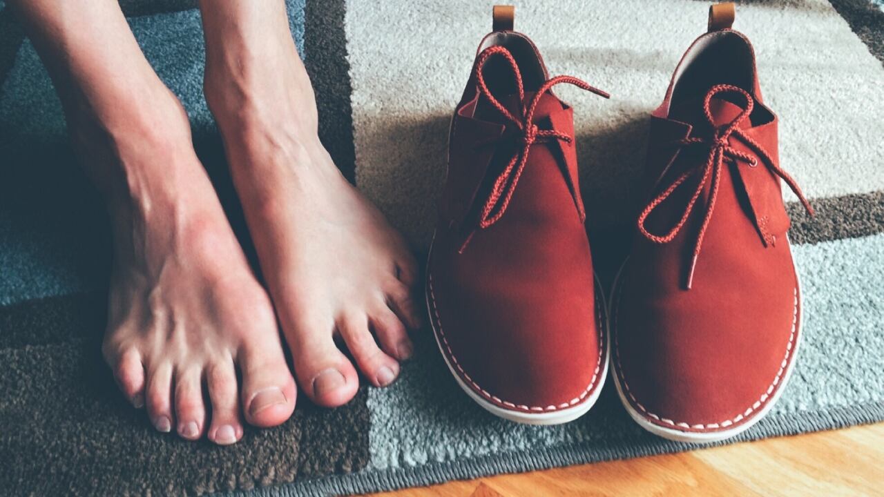 Un par de pies desnudos están posados sobre una alfombra y al lado de unos zapatos de gamuza rojos