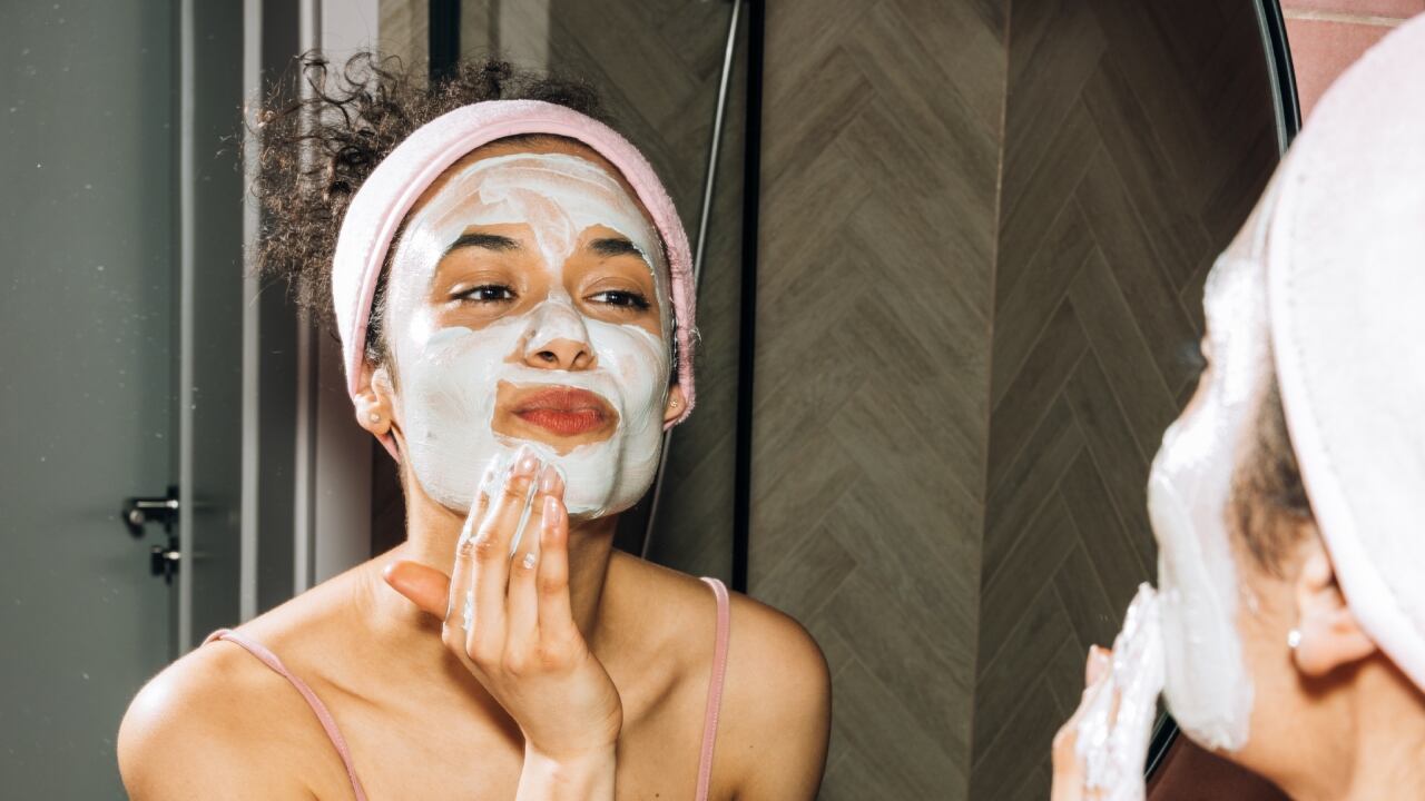 Mujer joven con una mascarilla facial está mirándose al espejo. Foto interior.