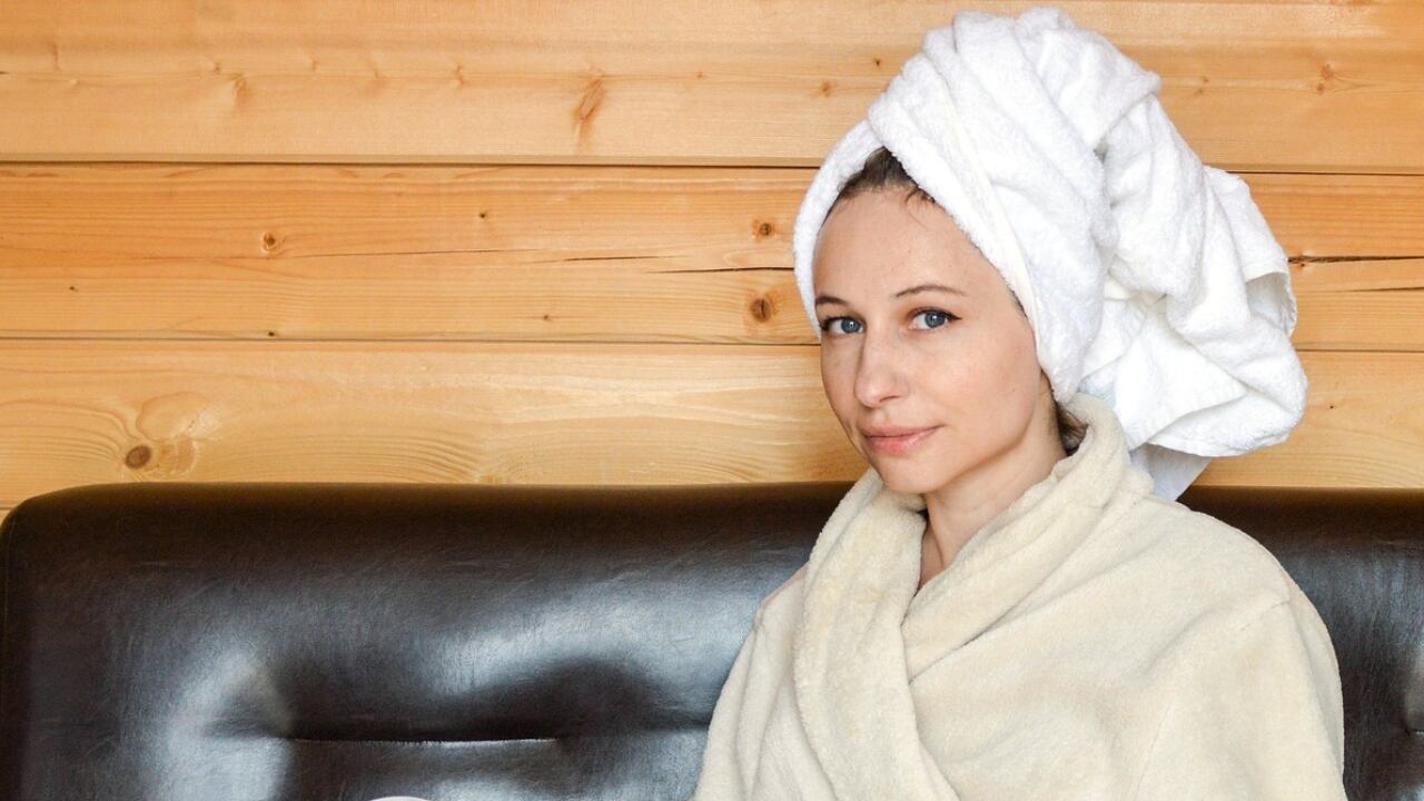 Mujer joven que parece recién salida de la ducha, con una toalla en la cabeza y una revista entre manos está sentada en un sillón de cuero negro.