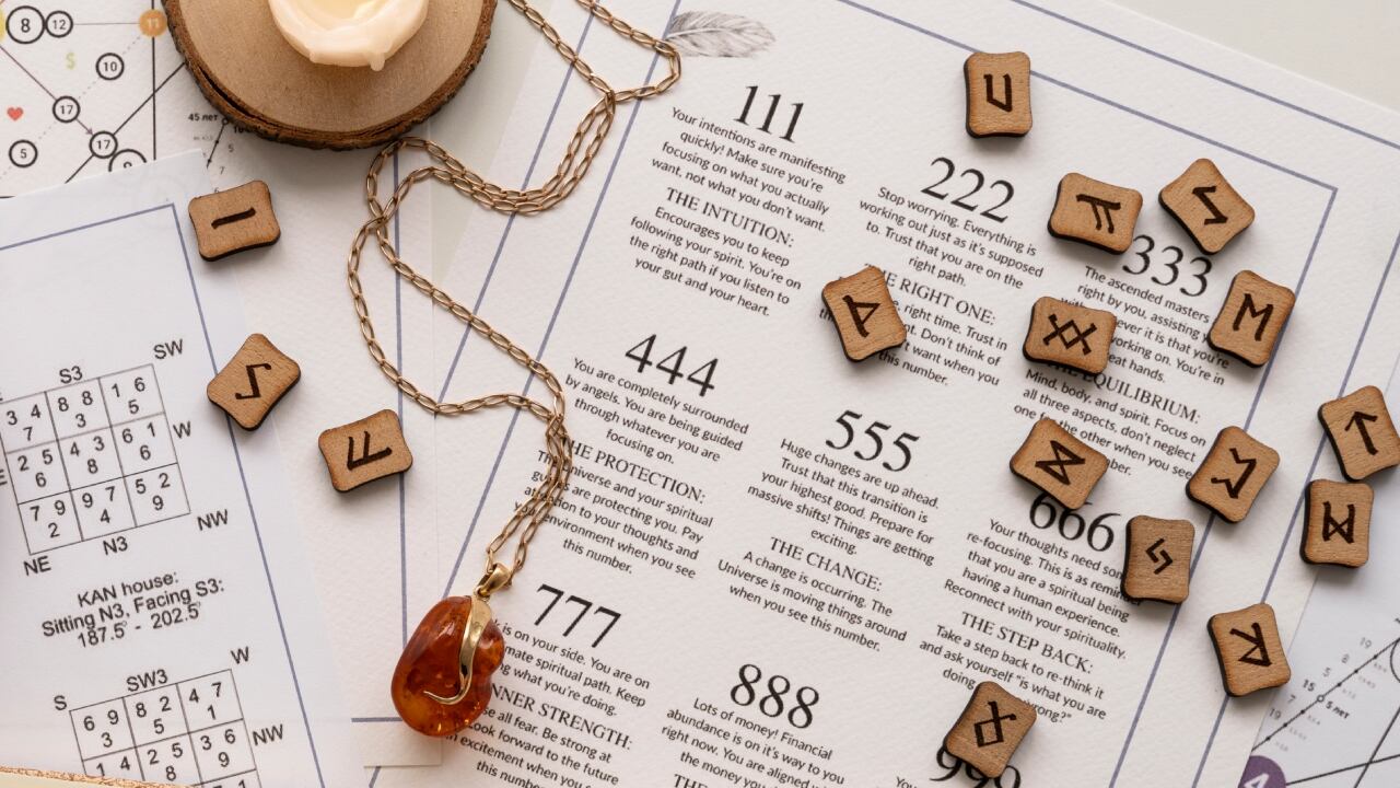 Runas de madera sobre una cartilla de numerología, al lado de una ágata que pende de una cadenita y una vela blanca encendida.