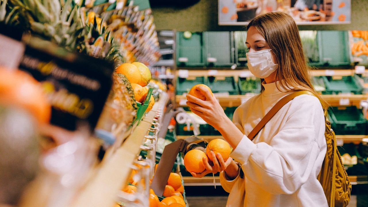 Una mujer joven vestida de blanco compra en el supermercado. Sostiene tres naranjas en sus manos.