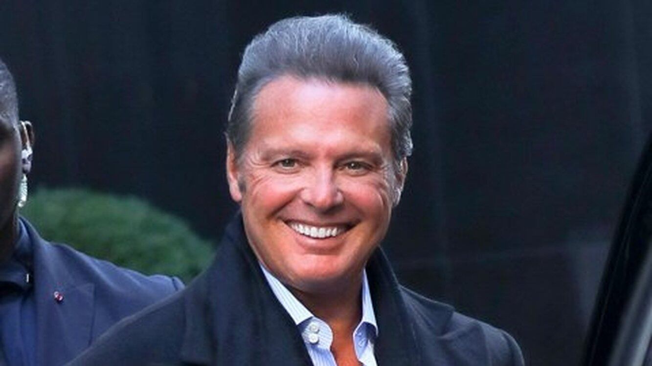 Hombre de avanzada edad sonriendo a la cámara con un abrigo y una bufanda negra y abajo tiene una camisa elegante abierta arriba