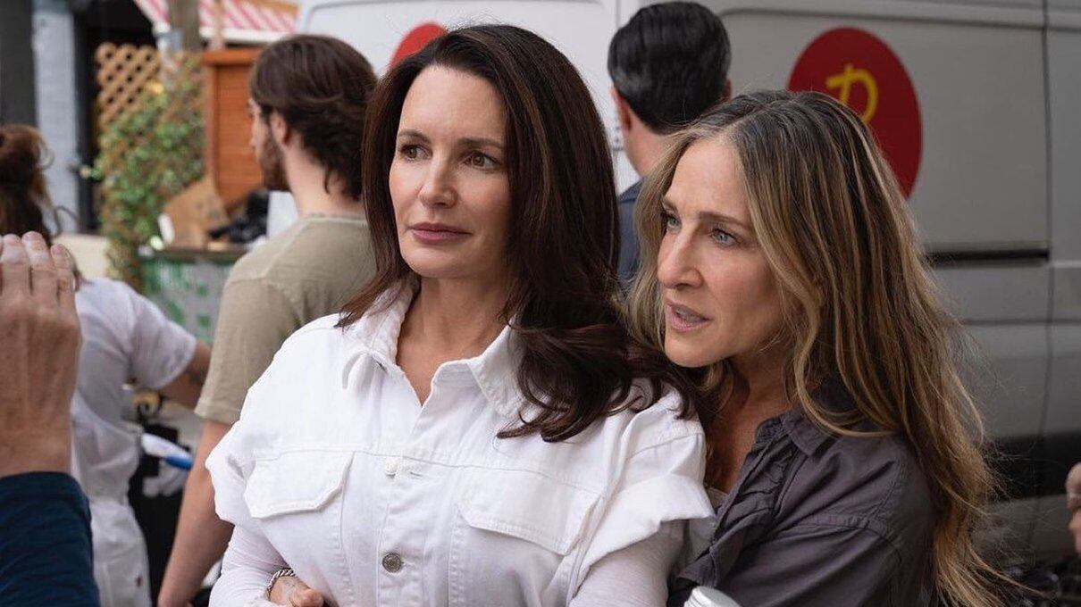 Kristin Davis, con una blusa blanca, es tomada por la cintura por Sarah Jessica, que va con una blusa gris oscuro, como deteniéndola, mientras están en una calle y se ven vehículos atrás.
