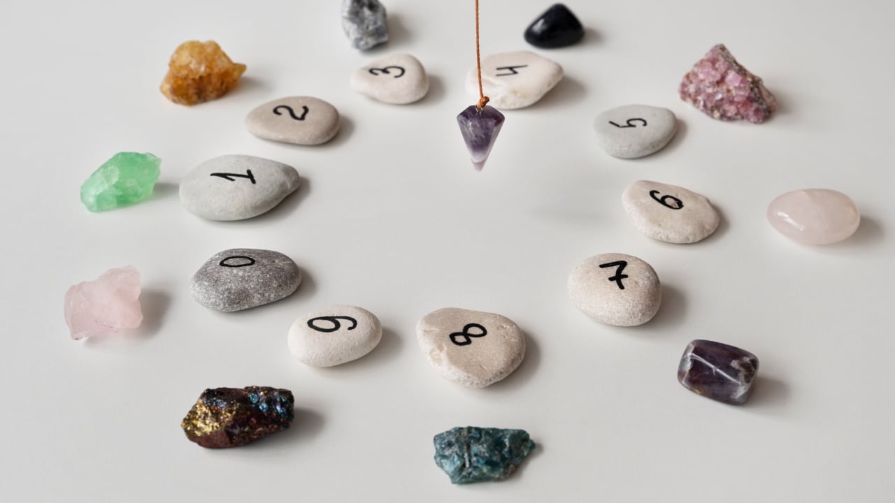 Dígitos dibugados sobre piedras dispuestas de forma circular. Al centro de ellas, pende un péndulo de amatista. Afuera de cada número, hay una piedra de un color diferente.