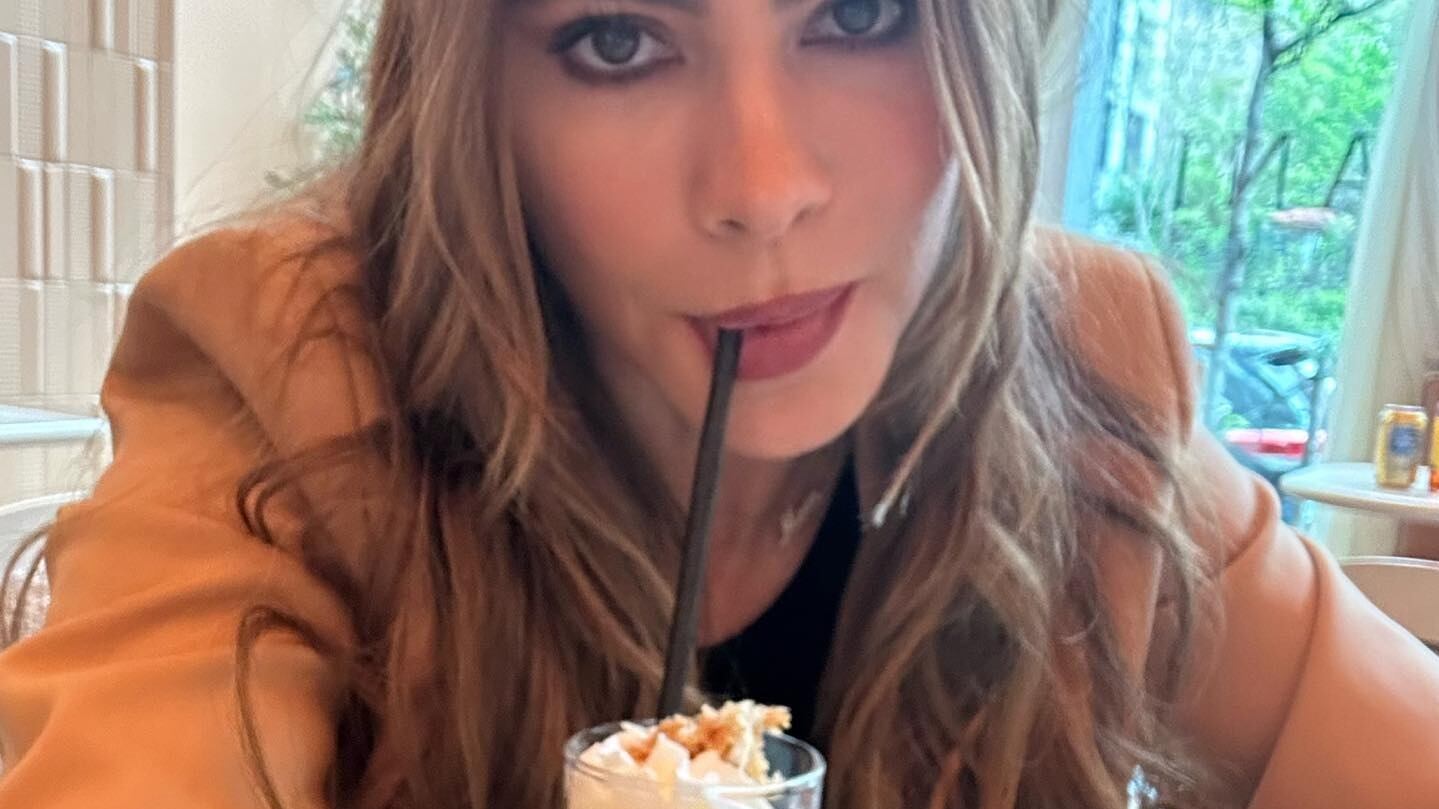 Mujer maquillada tomando una malteada con un popote en la boca, con chaqueta café y playera negra