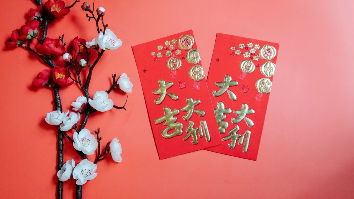 Dos ramas de cerezos en flor acompañan a dos tarjetas con caracteres y decoraciones chinas. Los cuatro elementos están sobre un fondo rojo pálido.